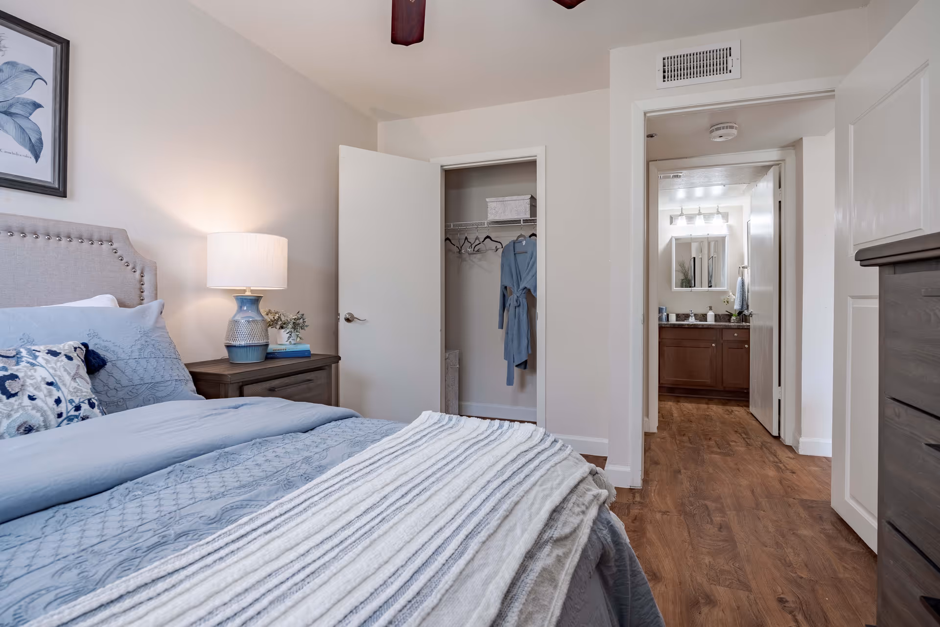 A cozy bedroom with a neatly made bed featuring blue bedding and a white textured throw blanket. A wooden nightstand with a blue and white lamp and a small plant sits beside the bed. An open closet door reveals a few hangers and a blue robe hanging inside. The room has wood flooring and light-colored walls. A doorway leads to a bathroom with a vanity, mirror, and lighting fixtures visible.