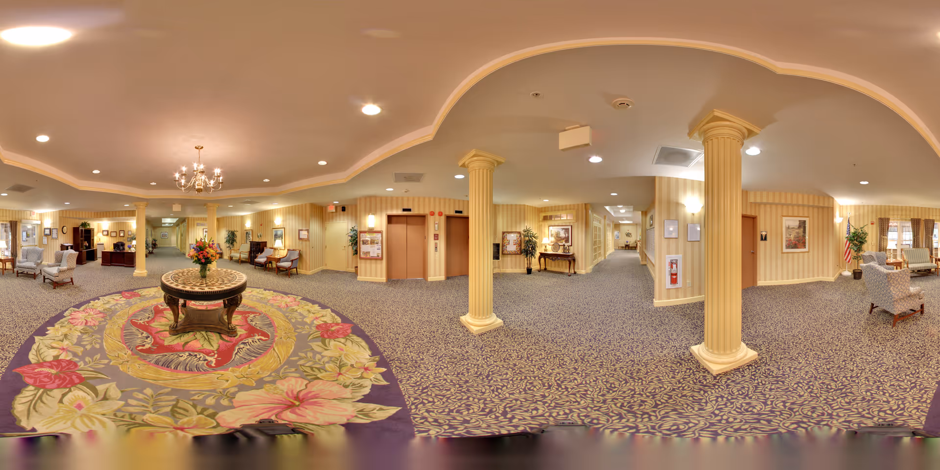 Spacious ornate senior living lobby with a central floral rug and table, columns, seating areas and elevators.