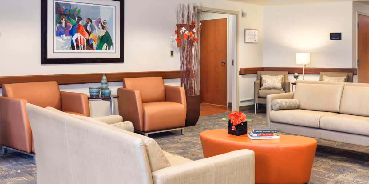 A bright, neatly furnished common lounge with beige sofas, orange armchairs, an orange ottoman coffee table, and wall artwork.