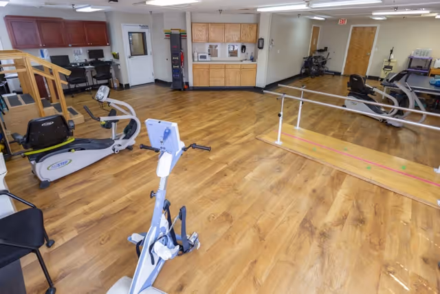 A spacious rehabilitation room with wooden flooring featuring exercise equipment including a stationary bike, a rowing machine, parallel bars for walking practice, and a kitchenette area with cabinets and a microwave. The room has white walls, ceiling lights, and a door leading to another area.
