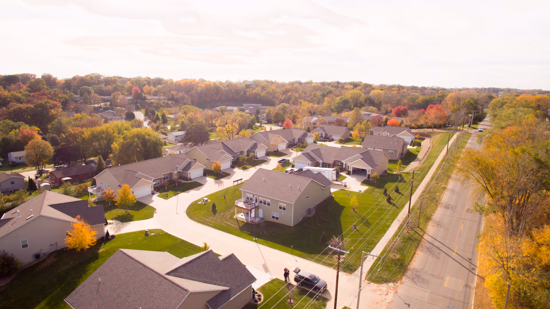 Aerial view of a residential community with multiple single-story houses surrounded by trees with autumn foliage. A road runs along the right side of the community, and the area is bathed in soft sunlight.