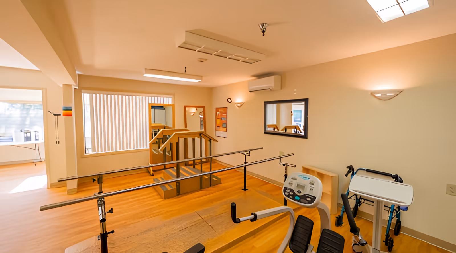 Interior view of a rehabilitation center room with wooden flooring, parallel bars for walking therapy, exercise equipment, mirrors on the walls, and a window with vertical blinds allowing natural light.