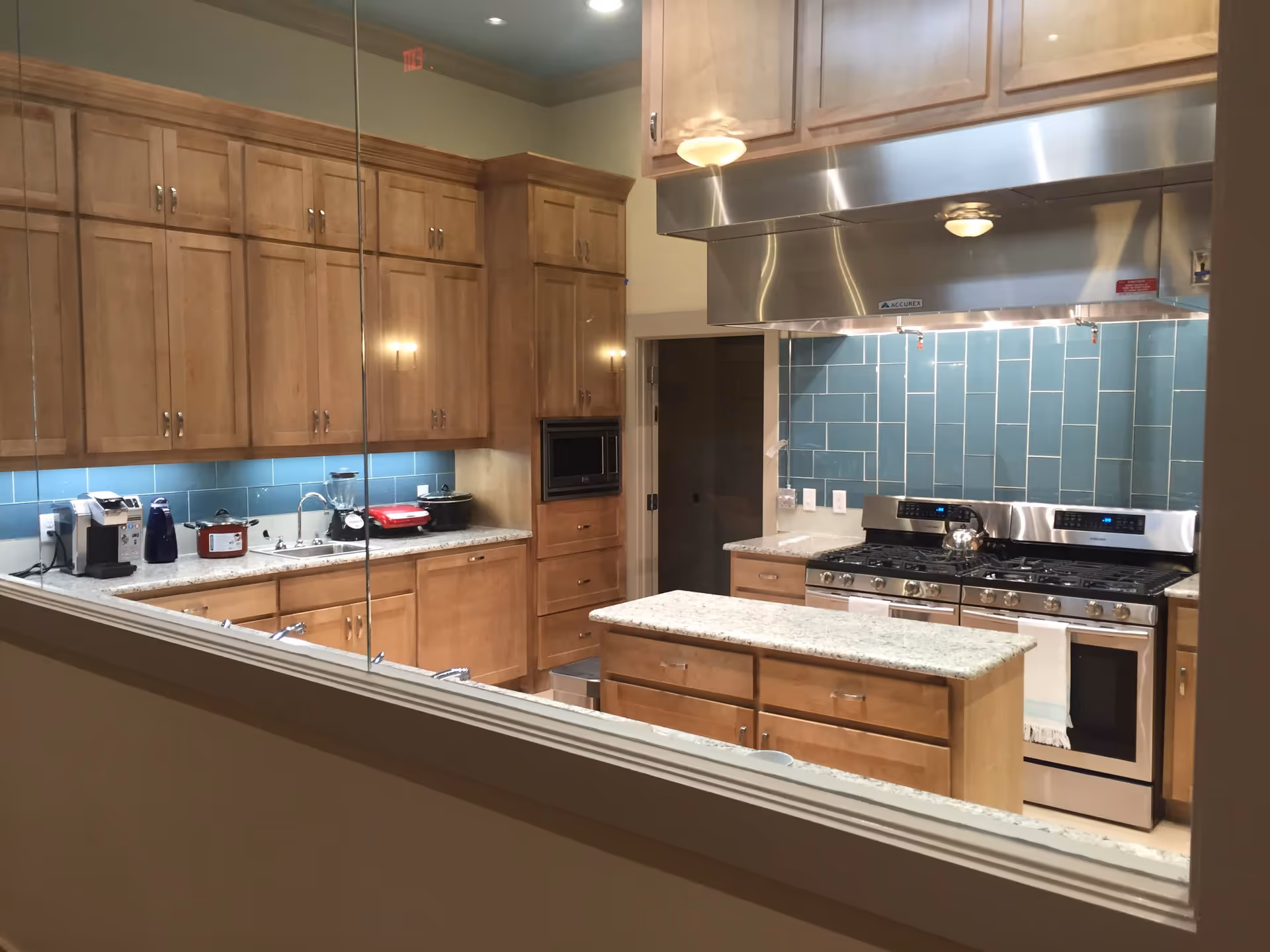 View through a glass window into a kitchen with wooden cabinets, a granite countertop island, a stainless steel stove with a hood, and a blue tiled backsplash. Various kitchen appliances are on the counter including a coffee maker, blender, and slow cooker.