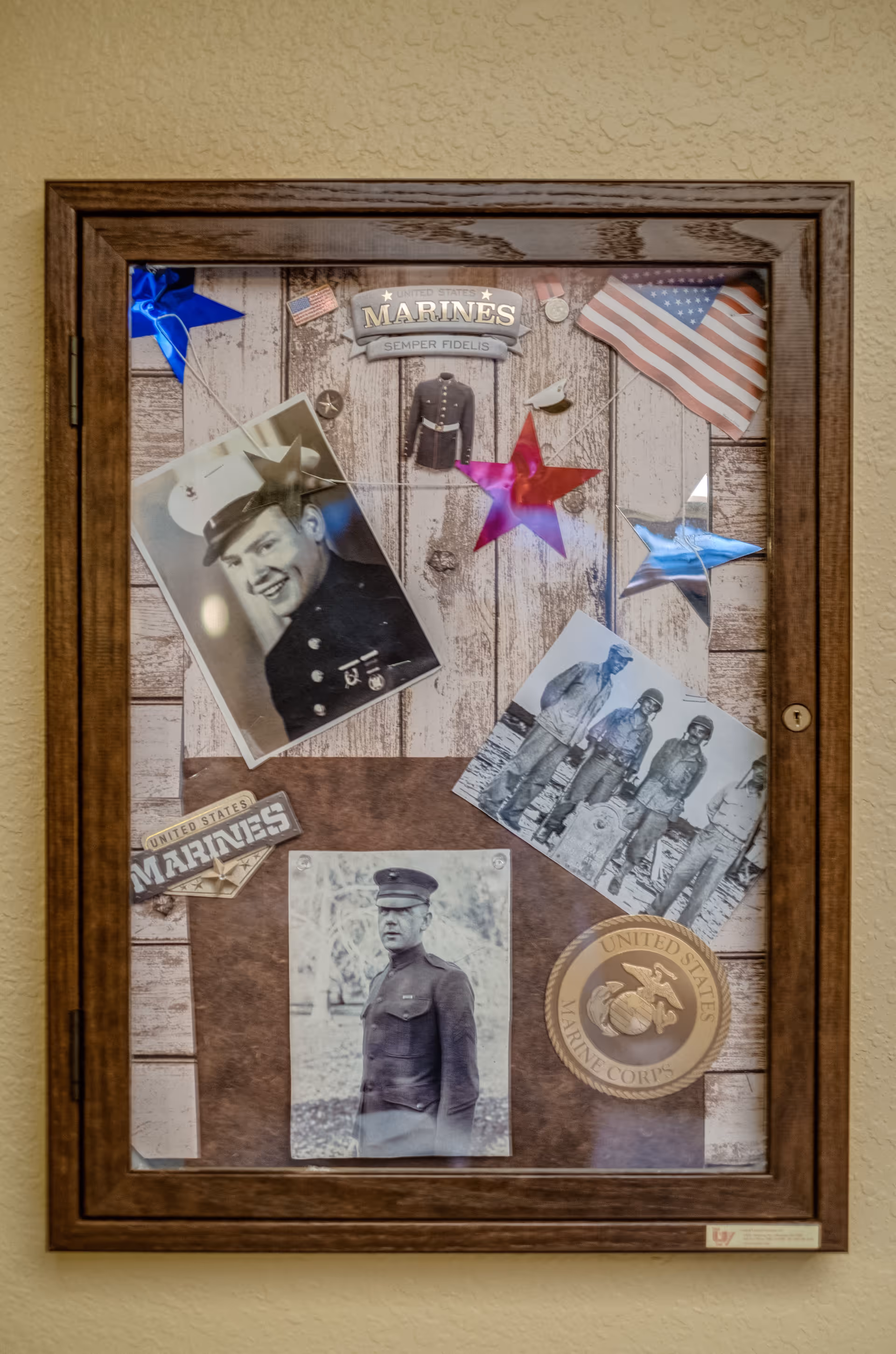 A wooden framed display case mounted on a wall containing vintage black and white photographs of U.S. Marines, a miniature Marine Corps dress uniform, American flags, colorful star decorations, and Marine Corps insignia and badges.