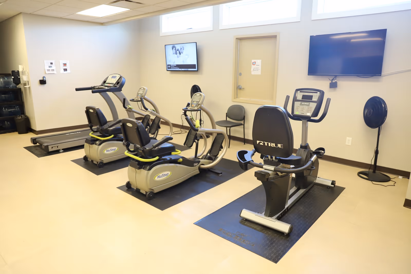A fitness room with exercise equipment including two NuStep recumbent cross trainers, a treadmill, and a TRUE recumbent exercise bike. The room has beige walls, a light-colored floor, two wall-mounted televisions, a fan, and a water dispenser in the corner.