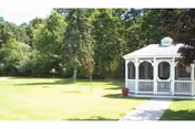 White gazebo beside a concrete path on a grassy lawn with trees in the background.