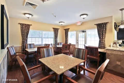 A bright dining room with multiple square tables and wooden chairs with striped cushions. The room has large windows with dark patterned curtains, light yellow walls, and ceiling lights. There is a counter area on the right side with a coffee maker and cups.