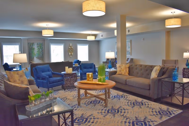 A spacious and well-lit living room with multiple seating options including gray sofas and blue armchairs arranged around a wooden coffee table on a patterned rug. The room features several lamps, modern ceiling lights, and large windows allowing natural light to enter. Decorative items such as plants, candles, and artwork adorn the space.