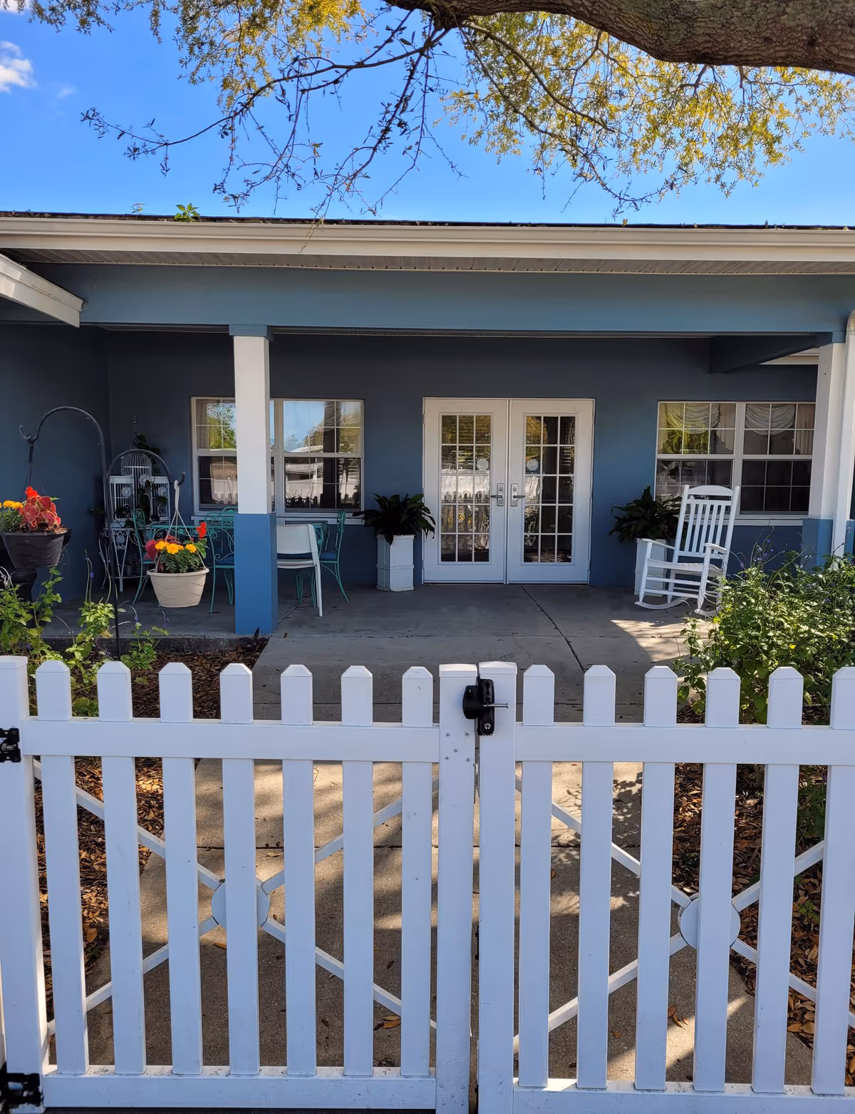 White picket gate opens onto a covered blue porch with French doors, potted plants, chairs and a white rocking chair.