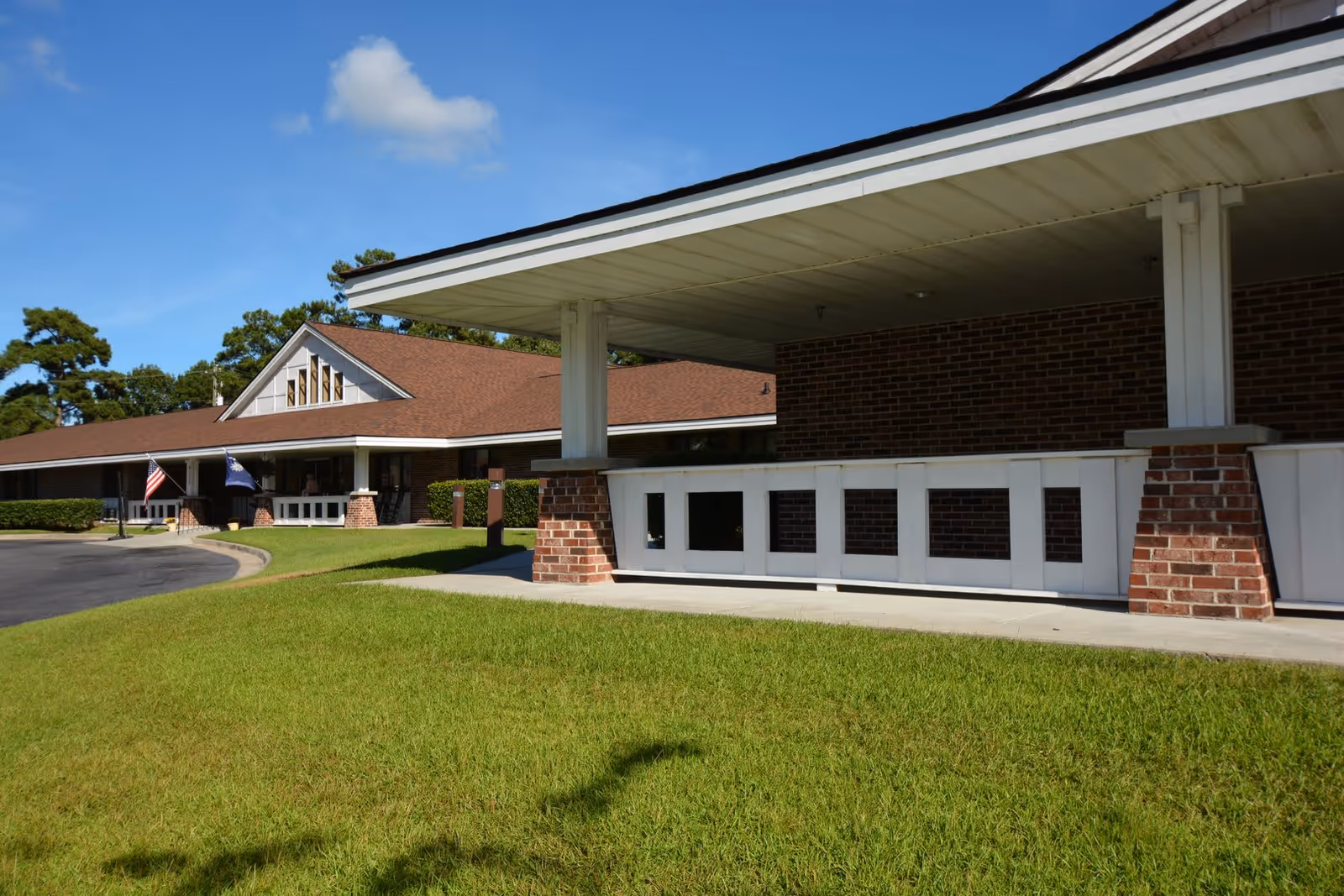 Exterior view of a single-story brick building with a brown shingled roof and white trim, surrounded by green grass and trees under a clear blue sky. Two flags are displayed near the entrance.