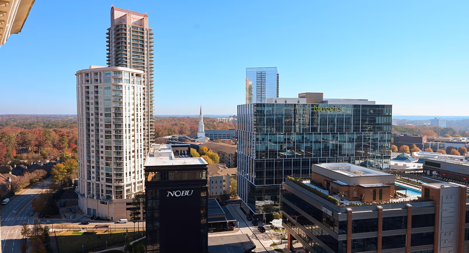 View of a cityscape featuring several modern high-rise buildings under a clear blue sky. The tallest building is a beige residential tower, with other commercial buildings including one with the sign 'NOBU' and another with 'Novelis'. Trees with autumn foliage are visible in the background.