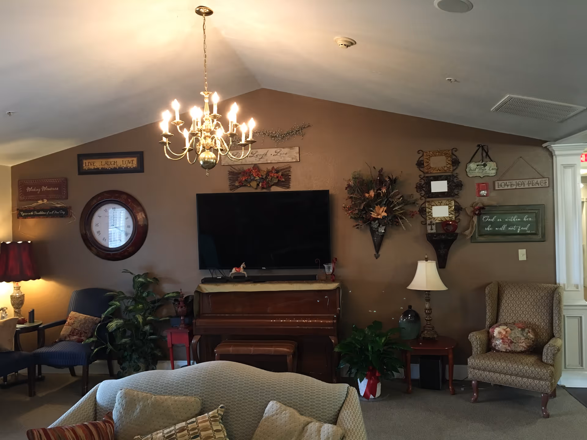 A cozy living room with a chandelier hanging from the ceiling, a wall-mounted flat screen TV above a piano, various framed inspirational signs and decorations on the brown wall, a red lamp on a side table, several armchairs, a couch with pillows, and indoor plants.