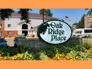 A landscaped entrance sign reading "Oak Ridge Place" with flowers, the facility building, and an American flag visible behind it.