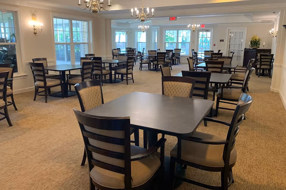 A spacious dining room with multiple square tables and cushioned chairs arranged neatly. The room has large windows allowing natural light to fill the space, beige carpet flooring, and elegant chandeliers hanging from the ceiling. The walls are painted white with classic wall sconces providing additional lighting.