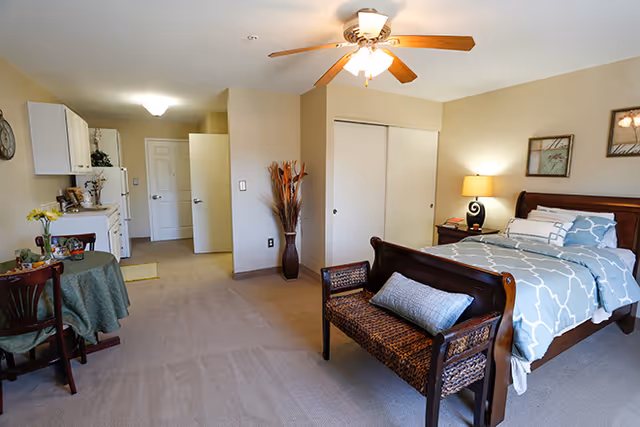 A spacious senior living bedroom with a wooden bed featuring blue and white patterned bedding, a wicker bench at the foot of the bed, a nightstand with a lamp, and framed artwork on the wall. To the left, there is a small dining table with two chairs and a kitchenette area with white cabinets. The room has beige walls, carpeted floor, and a ceiling fan with lights.