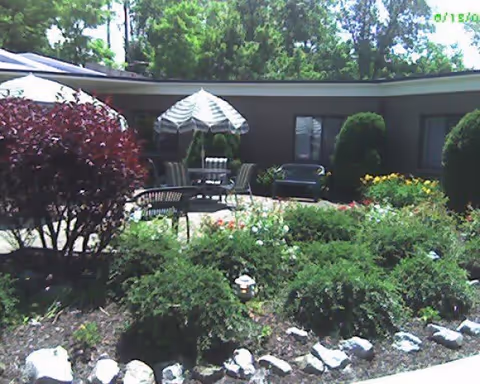 Outdoor garden area with various green shrubs, flowering plants, and a small tree with red leaves. There is a patio with a table, chairs, and a striped umbrella. The background shows a single-story building with windows and some bushes.