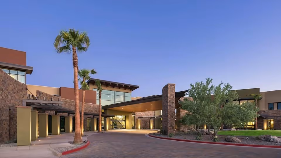 Exterior view of Clearwater Ahwatukee senior living facility at dusk, featuring modern architecture with stone and stucco walls, large windows, palm trees, and a covered entrance driveway.