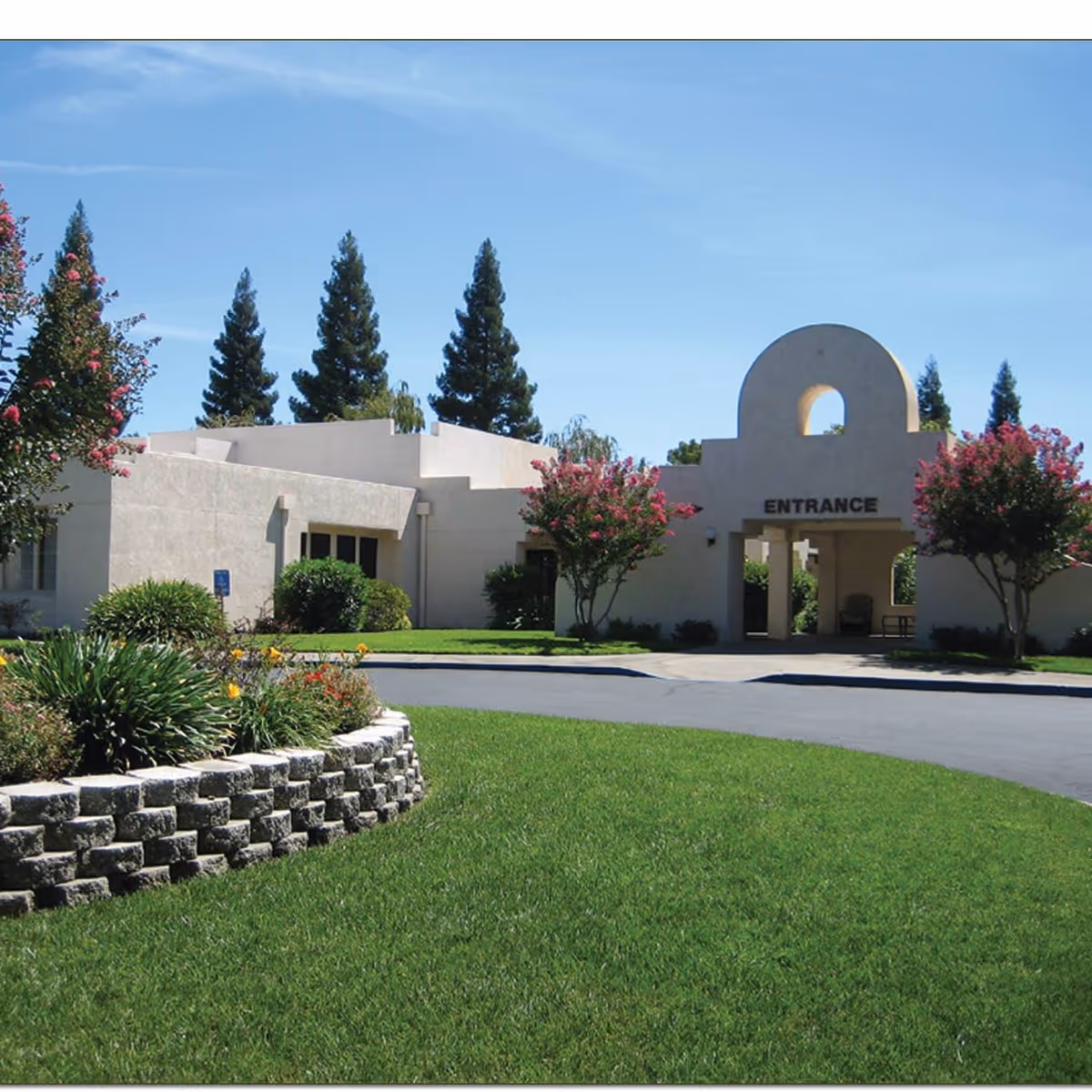 Front entrance of a low-rise stucco building with an arched entry labeled "ENTRANCE", surrounded by manicured lawn and trees.