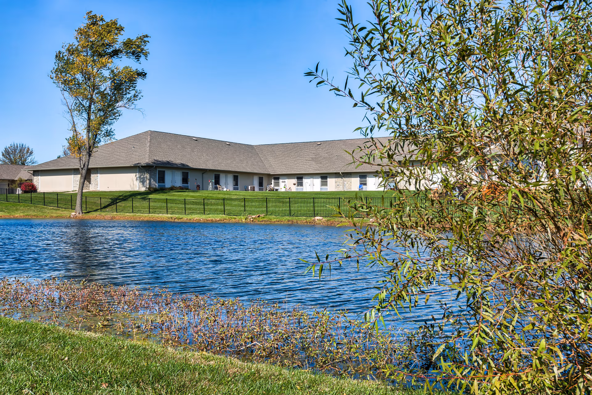 View of a single-story assisted living facility building behind a pond with clear blue water, surrounded by green grass and trees under a clear blue sky.