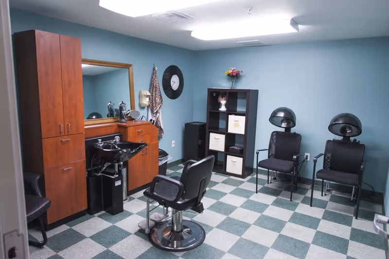 Interior of a senior living facility hair salon with teal walls and checkered green and white floor tiles. The room contains a wooden cabinet with a mirror and a black sink, a black salon chair in front of the sink, two black chairs with hair dryers above them, a black shelving unit with storage bins and a mannequin head, a clock on the wall, and a vase with flowers on top of the shelving unit.