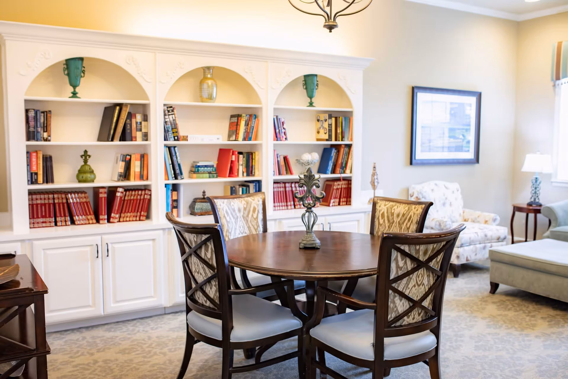 A cozy living room area with a round wooden table surrounded by four upholstered chairs. Behind the table is a white built-in bookshelf filled with books and decorative items. The room features a patterned carpet, a framed picture on the wall, a floral armchair, a side table with a lamp, and a large window with striped curtains letting in natural light.