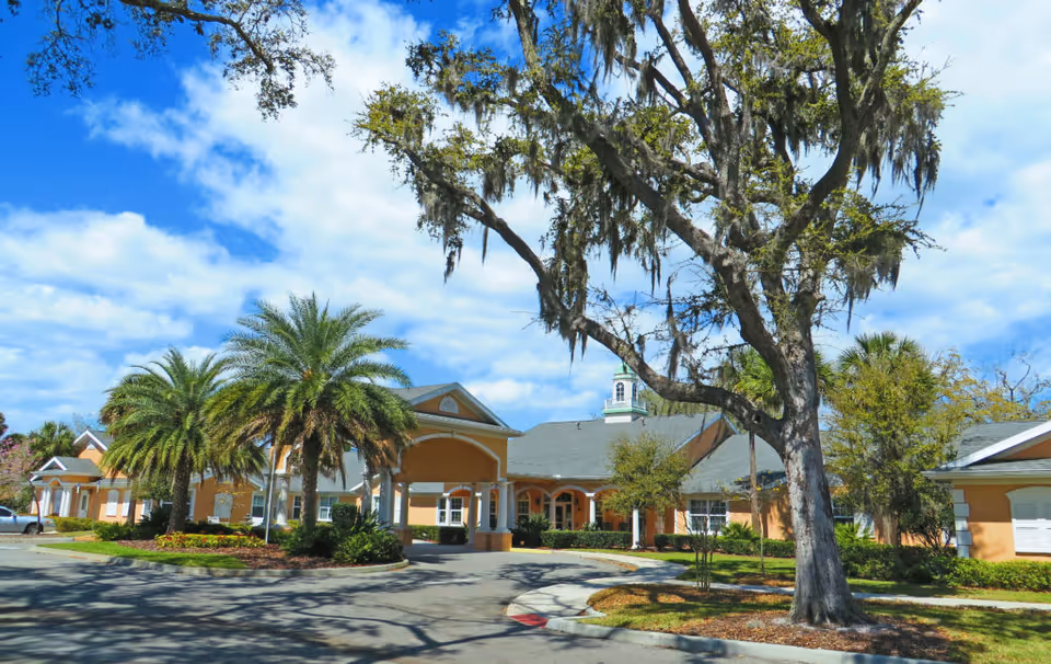 Exterior view of Addington Place of Titusville, showing a single-story building with a covered entrance, surrounded by palm trees and other greenery under a partly cloudy blue sky.