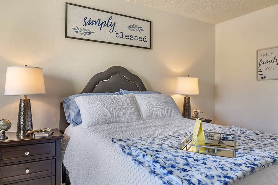 A cozy bedroom with a neatly made bed featuring white and blue bedding. There are two bedside tables with lamps on each side of the bed. Above the bed, a framed sign reads 'simply blessed'. Another framed sign on the adjacent wall reads 'Faith family love'.