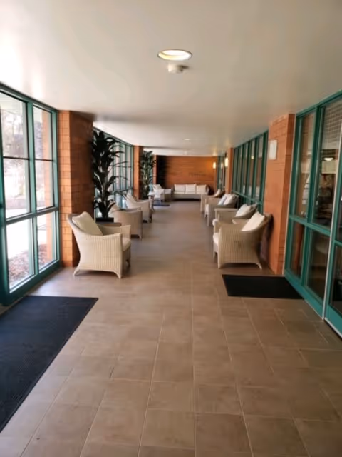 A long indoor corridor with large windows on the left and brick walls on the right. The corridor is furnished with multiple wicker armchairs with cushions arranged in pairs along the walls. There are also some potted plants and a seating area with a sofa at the far end.