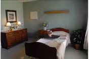 A neatly arranged bedroom in a senior living facility featuring a single bed with a beige blanket, a wooden headboard and footboard, a wooden dresser with two lamps and framed pictures on the wall, a small bedside table with a flower vase, and a potted plant near a window with curtains.