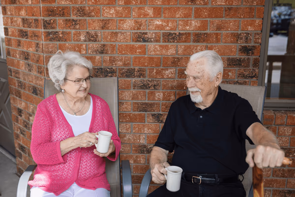 An elderly woman wearing a pink cardigan and an elderly man in a black shirt sit outside on chairs against a brick wall, each holding a white mug and smiling at each other.
