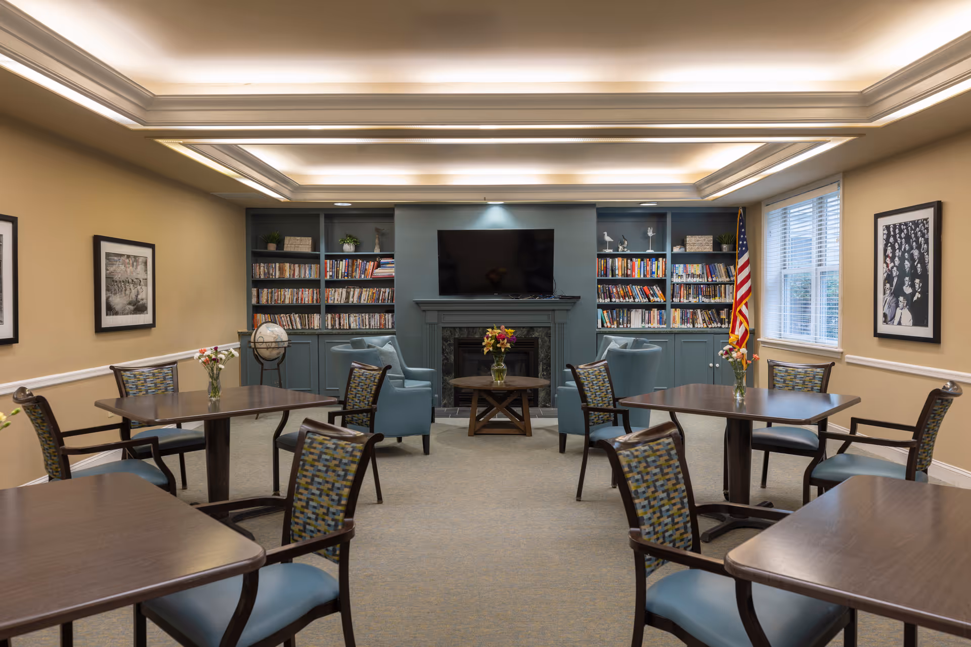 A well-lit common room with several tables and chairs arranged for seating. The room features a fireplace with a TV mounted above it, flanked by built-in bookshelves filled with books and decorative items. There are framed black and white photographs on the walls, a globe on a stand, and an American flag near a window with blinds. Fresh flowers in vases are placed on the tables and a small coffee table in front of the fireplace.