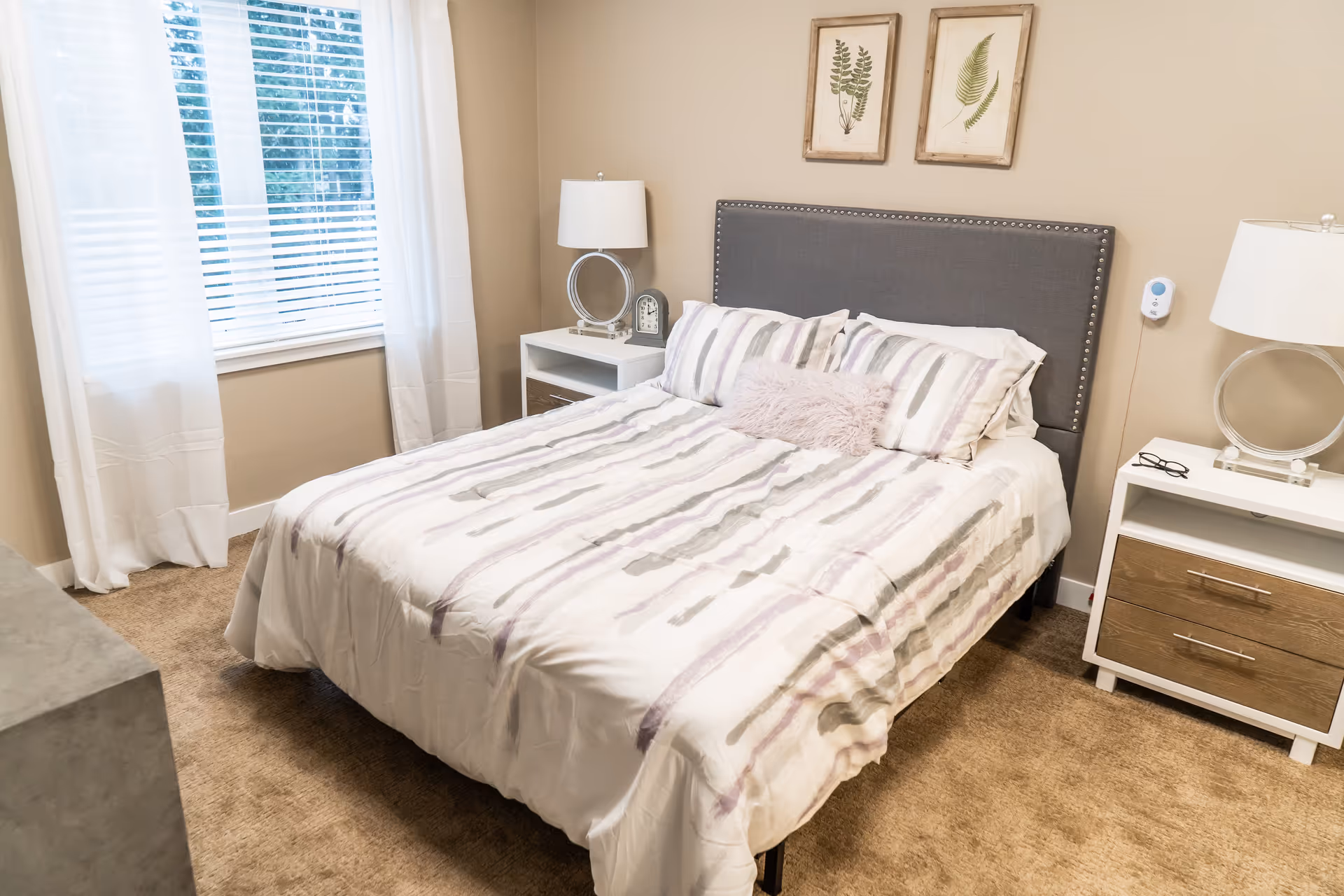A neatly made bedroom with a double bed, gray upholstered headboard, nightstands with lamps, and a window with white curtains.