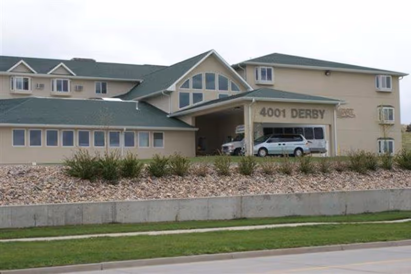 Exterior view of a beige senior living facility building with green roofs, multiple windows, and a covered entrance area with vehicles parked underneath. The address number 4001 DERBY is visible on the building.