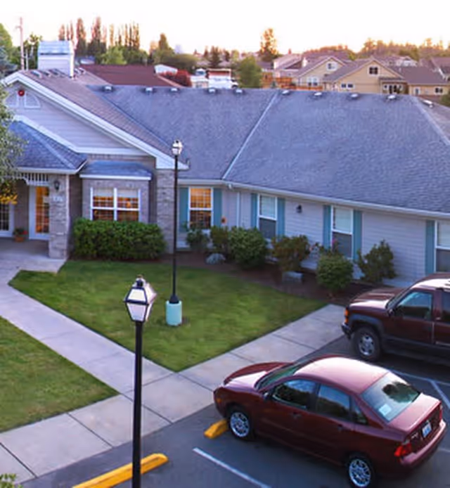 Exterior view of a single-story residential building with a gray roof and light-colored siding, surrounded by green bushes and a well-maintained lawn. There are two cars parked in the parking lot adjacent to the building, and a sidewalk with street lamps runs through the grassy area.