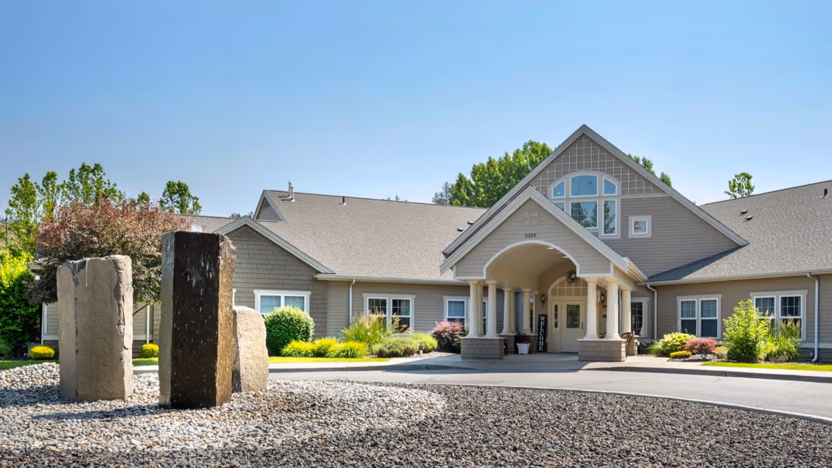 Exterior view of a single-story senior living facility building with a peaked roof and a covered entrance supported by columns. In front of the building is a circular driveway with a rock garden featuring several large standing stones. The landscaping includes green shrubs and trees under a clear blue sky.