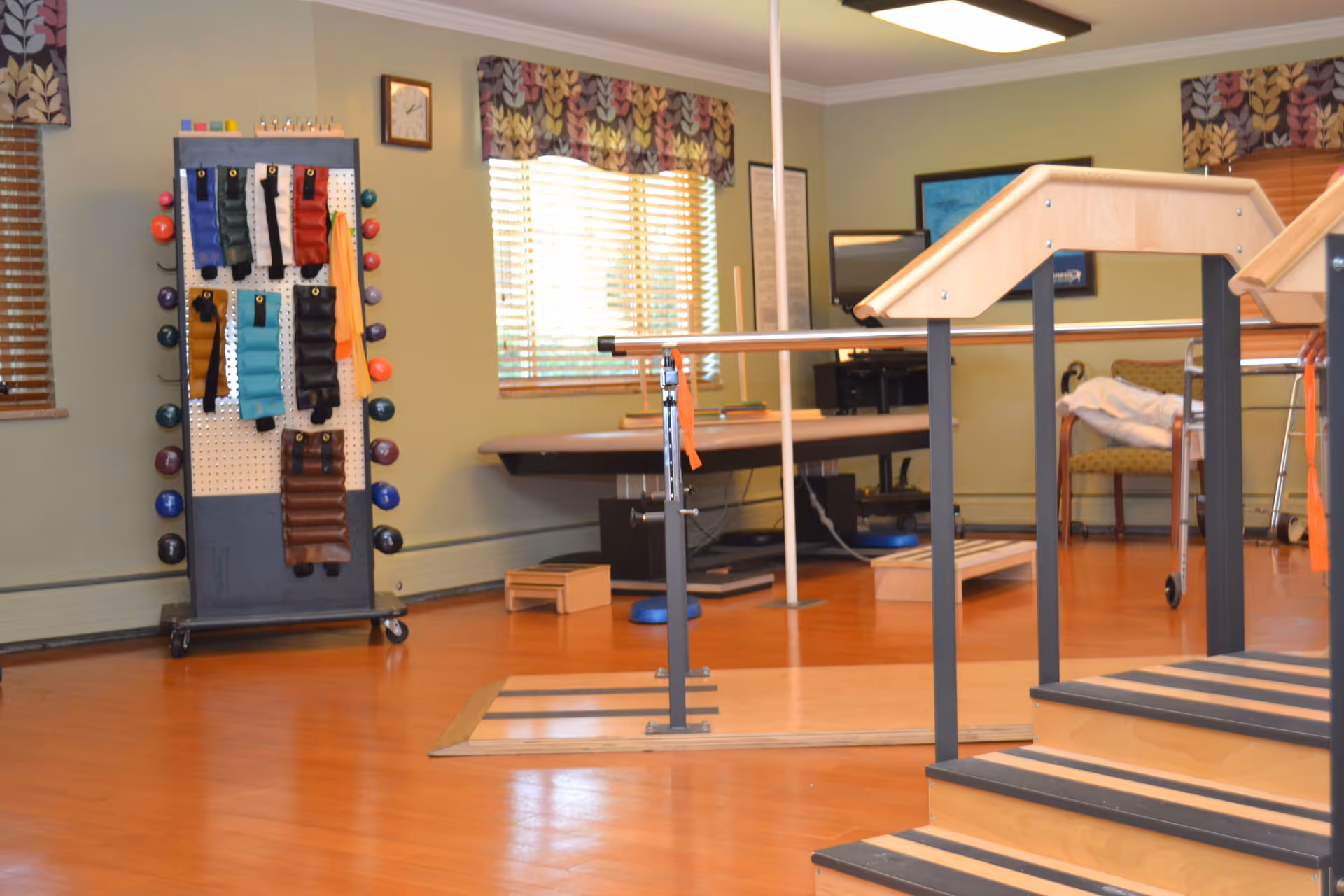 A physical therapy room with wooden stairs equipped with handrails for rehabilitation exercises. The room has hardwood floors, windows with patterned valances, and various therapy equipment including weighted bags hanging on a stand and parallel bars. There is a chair and a walker in the background.
