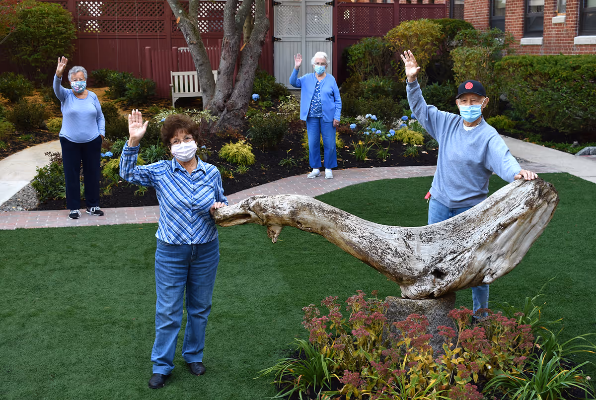 Four elderly individuals wearing face masks standing outdoors on a lawn with a large piece of driftwood sculpture in the center. They are spaced apart and waving at the camera. The background shows a garden with plants, a tree, and a red fence with a brick building.