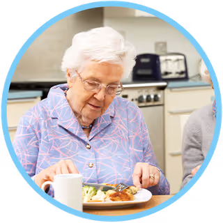 An elderly woman seated at a table eating a meal in a kitchen-like dining area.