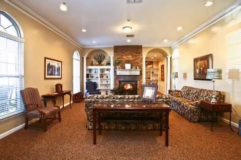Well-lit living room with patterned sofas and chairs facing a lit stone fireplace flanked by built-in bookshelves and end tables.