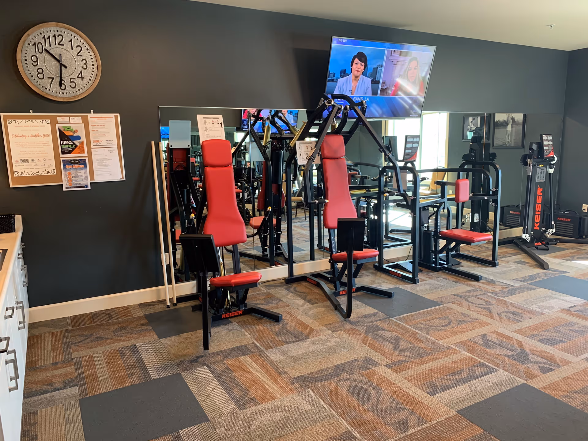 Fitness room with exercise machines featuring red seats and black frames, a large wall clock, bulletin board with notices, a wall-mounted TV showing a news program, and a mirrored wall reflecting the equipment.