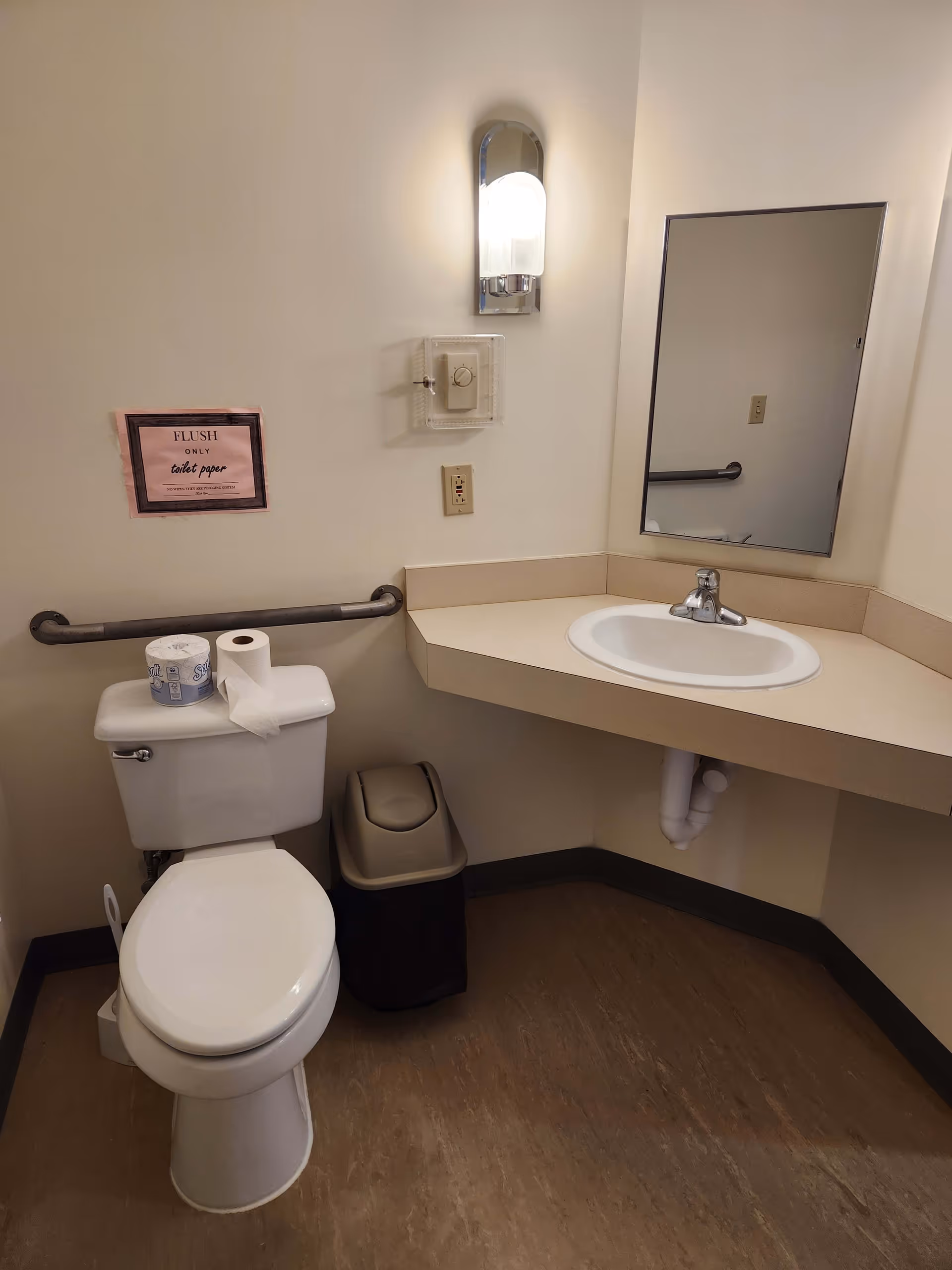 A small bathroom with a white toilet, a roll of toilet paper on top of the tank, a beige trash can next to the toilet, a beige countertop with a white sink, a wall-mounted light fixture above the sink, a rectangular mirror, and a grab bar on the wall behind the toilet. There is a sign on the wall that reads 'FLUSH ONLY toilet paper.'