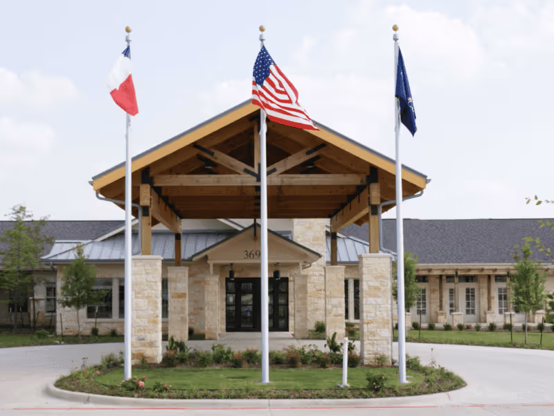 Front exterior view of a senior living facility named Ellery Arbor with a covered entrance supported by wooden beams and stone pillars. Three flagpoles stand in front, displaying the Texas state flag, the United States flag, and another flag. The building has a stone facade and a gray roof, with some landscaping including small trees and bushes.