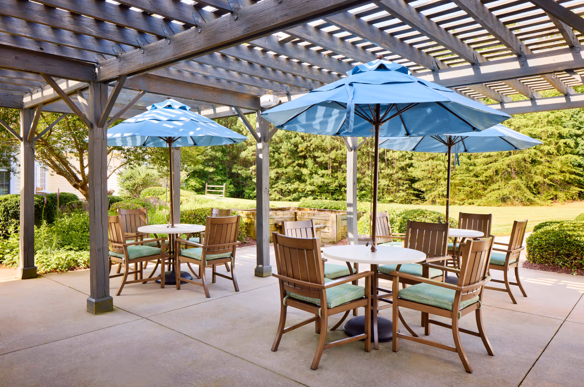 Outdoor patio area with wooden pergola overhead, featuring round tables with blue umbrellas and wooden chairs with green cushions, surrounded by greenery and trees.