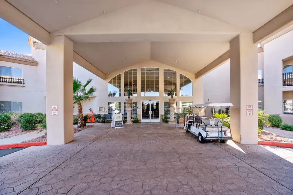 Entrance area of The Montecito Senior Living facility with a covered driveway, two large columns, potted plants, a golf cart parked on the right side, and glass doors leading inside. There are signs indicating no parking in the fire lane and a sign advertising daily tours.