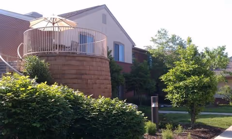 Raised circular patio with an umbrella and railing attached to a brick building, surrounded by shrubs and trees.