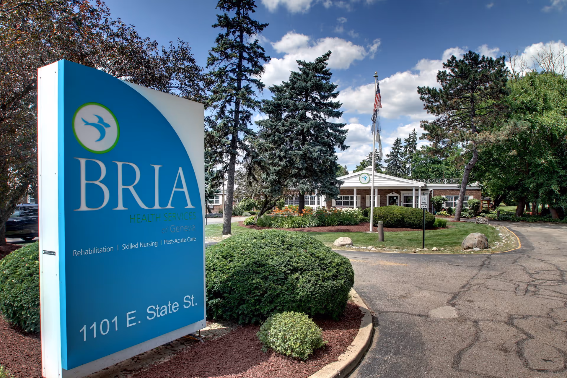 Exterior view of BRIA Health Services of Geneva, showing a large blue sign with the facility name and address 1101 E. State St. The building is surrounded by trees, bushes, and a curved driveway under a partly cloudy sky.