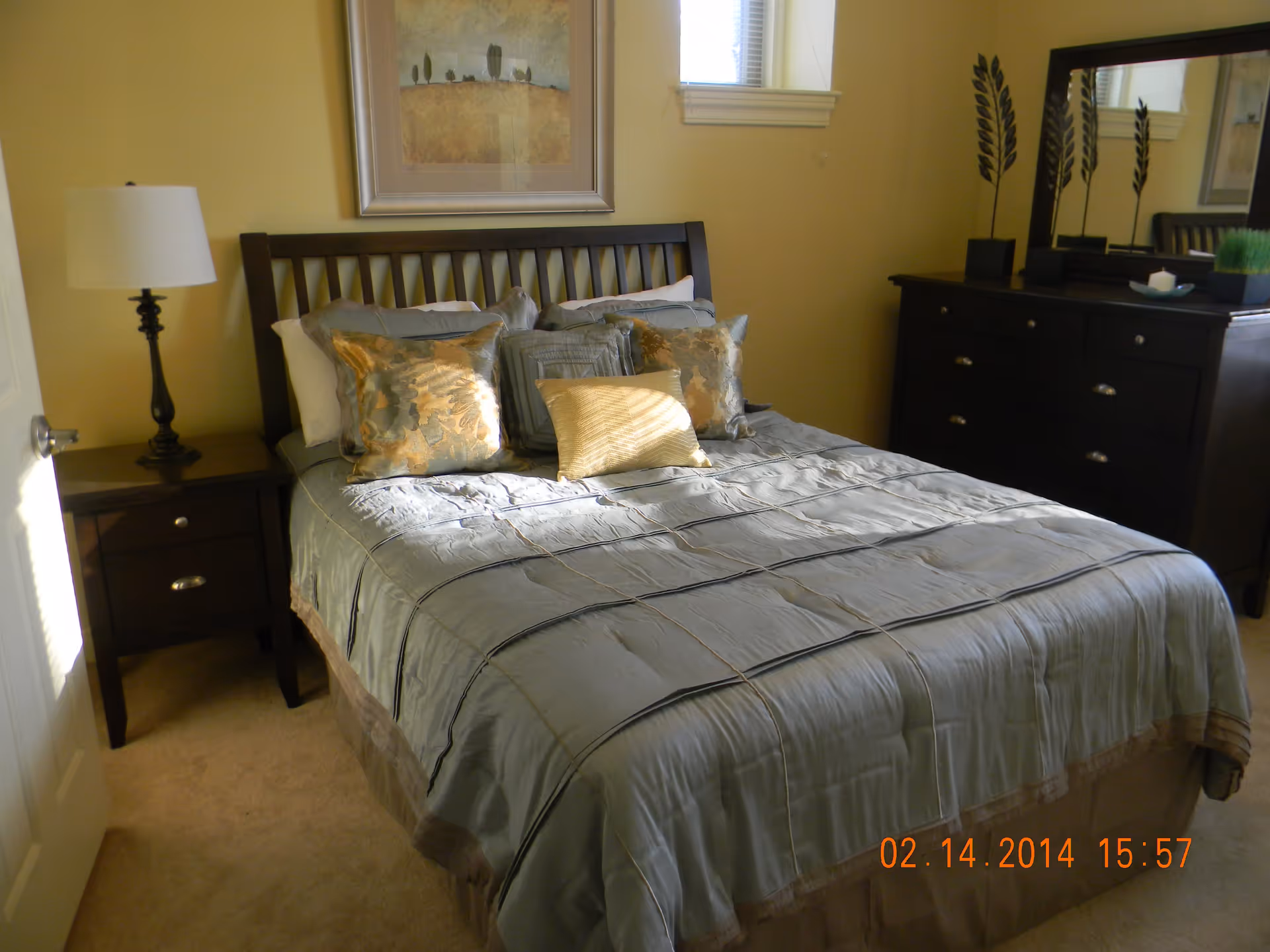 A neatly made bed with a wooden headboard in a bedroom. The bed has a light blue comforter and several decorative pillows in shades of gold and blue. To the left of the bed is a dark wooden nightstand with a lamp. On the right side of the room is a dark wooden dresser with a mirror above it and decorative items on top. A framed picture hangs on the wall above the bed, and a small window is visible on the right wall.