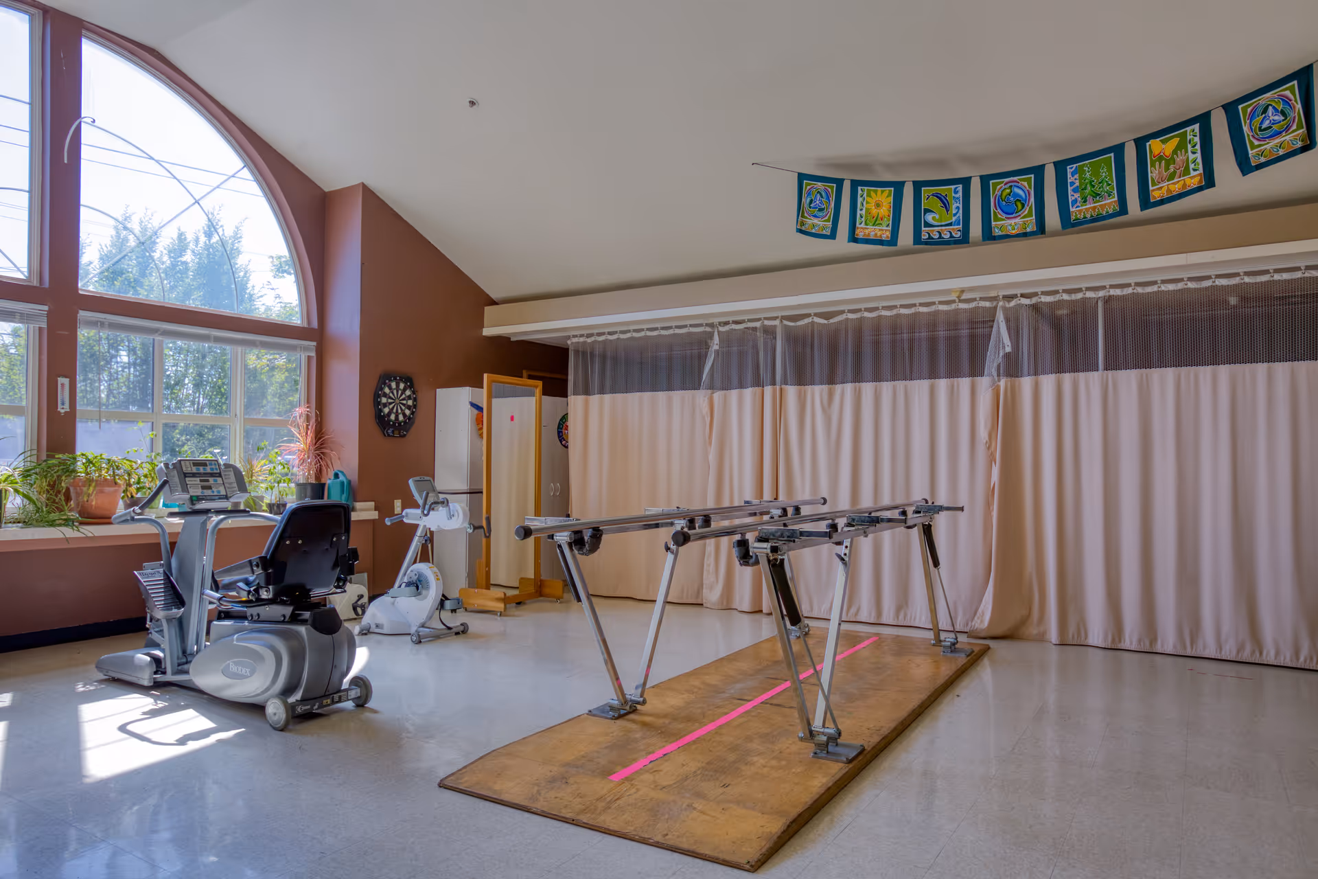 A spacious rehabilitation room with large arched windows letting in natural light. The room contains exercise equipment including a recumbent bike and a stationary bike, a wooden parallel bar setup for walking exercises, a dartboard on the wall, and a row of colorful decorative flags hanging near the ceiling. There are plants on the windowsill and a beige curtain partition in the background.