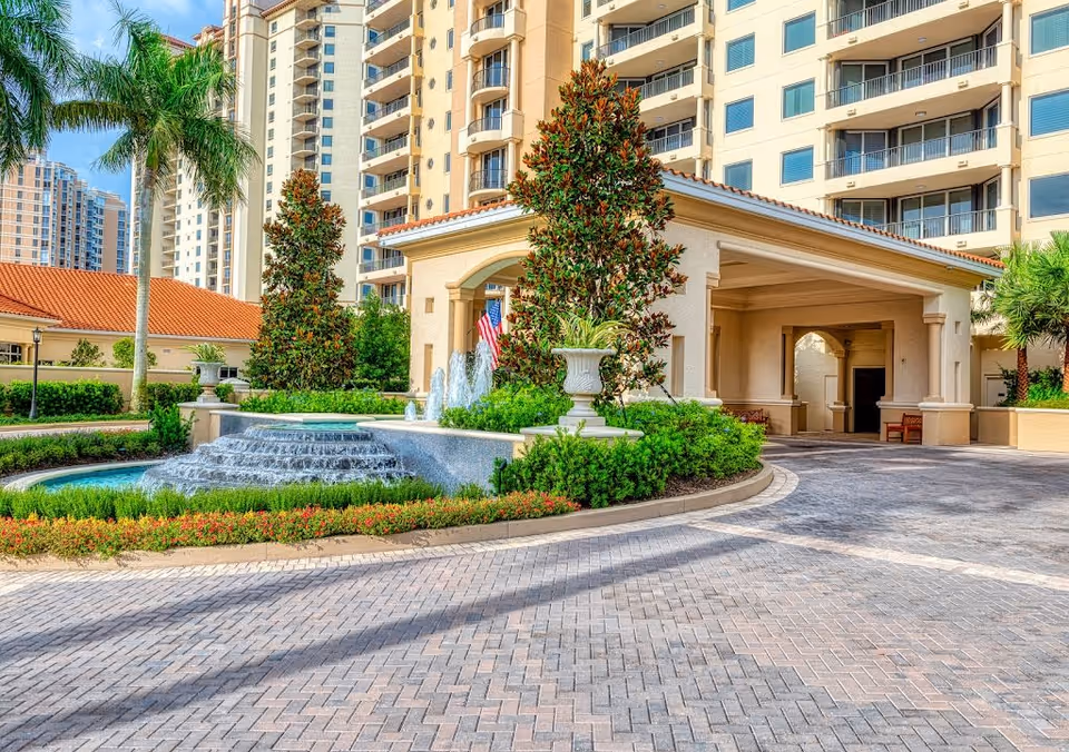Entrance area of The Marbella at Pelican Bay featuring a covered drop-off zone, a tiered water fountain surrounded by greenery and flowers, palm trees, and a multi-story residential building in the background under a blue sky.