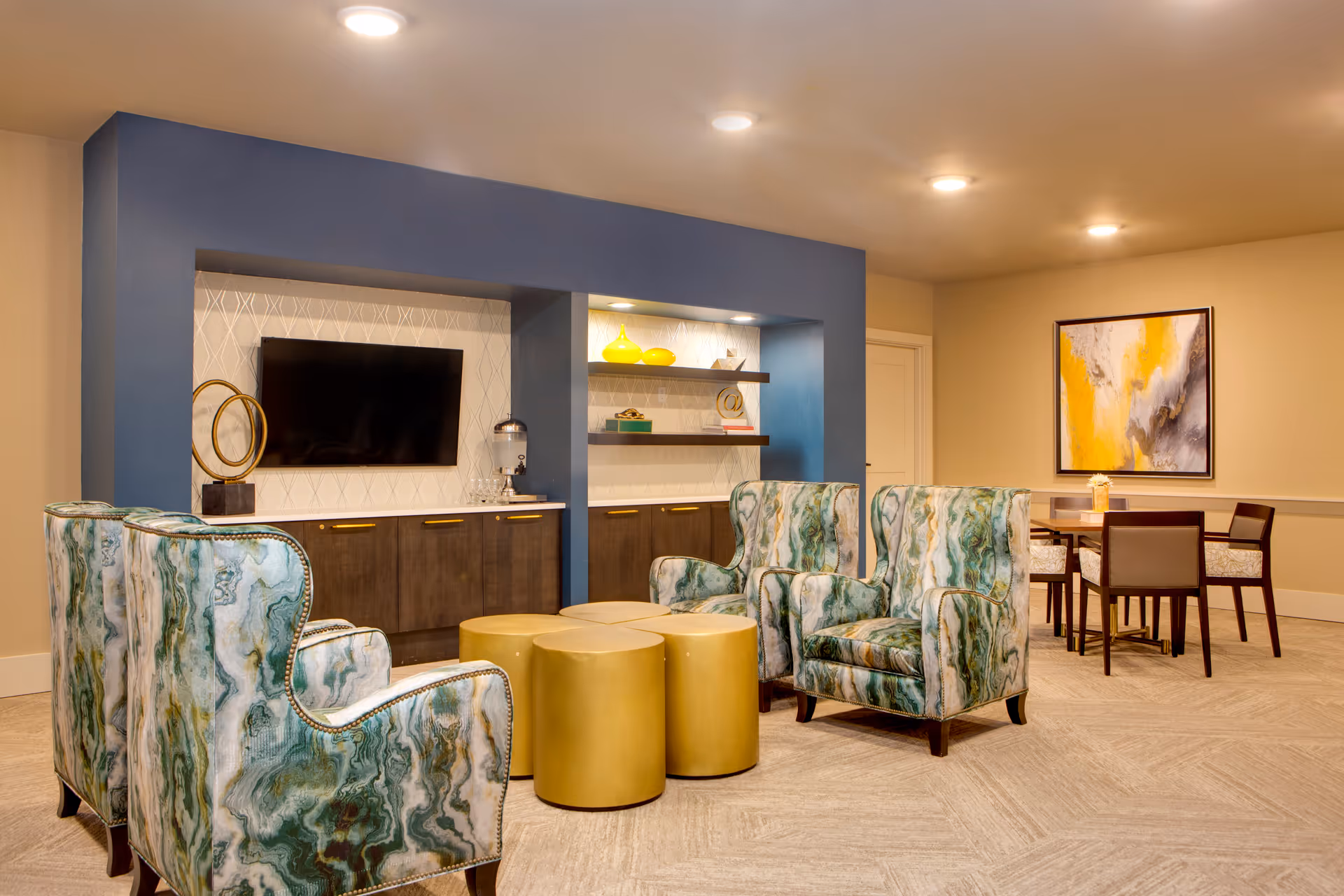 A cozy senior living common area with four patterned armchairs arranged around three gold cylindrical ottomans. A built-in entertainment center with a flat-screen TV, decorative items, and shelves is set against a blue accent wall. In the background, there is a dining table with four chairs and a large abstract painting with yellow, white, and gray tones on the wall.
